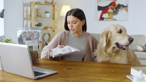 Medium shot of middle-aged Caucasian woman sitting with her dog in front of laptop screen, and discussing dog food with her friend by video link
