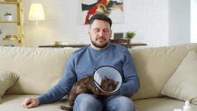 Lockdown of man sitting on couch with post-surgical cat in funnel cone on his knees, petting it, listening to doctor and nodding