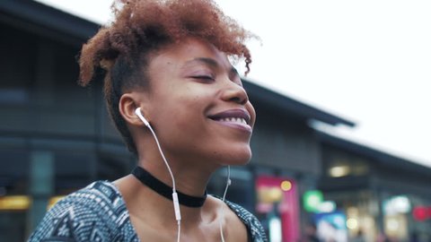 Slow motion close up of happy young African woman enjoying music dancing in the street. Joyful black young woman listening to music wear earphone looking at camera smile at Asian urban background. 