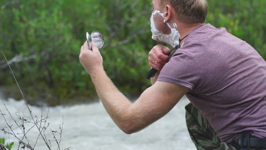a young man shaves his face with a large sharp knife, looks into a small round mirror, being at a mountain river