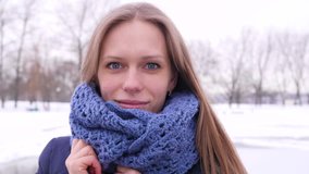 pretty blue-eyed woman in blue knitted scarf at the winter city park who looking at the camera and corrects her long hair in 4K slow motion close up video.