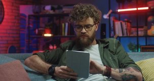 Caucasian young stylish man in glasses sitting on the sofa in the cozy living room at night, watching or reading something on the tablet device, scrolling and tapping.