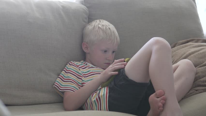 The boy sits on the couch and plays in the tablet. He moves his finger on the glass of the tablet.