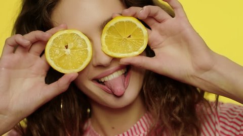 Playful girl having fun with lemon slices on yellow background. Cheerful woman holding two lemon slices in front of eyes. Portrait happy woman showing tongue in studio. Healthy food and diet concept