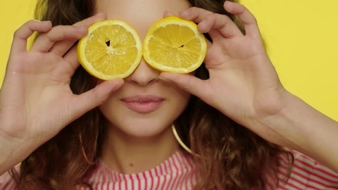 Young woman kissing lips with lemon slices in front of eyes on yellow background. Cheerful woman posing with orange slices in front of face in studio. Fashion model having fun with lemon halves