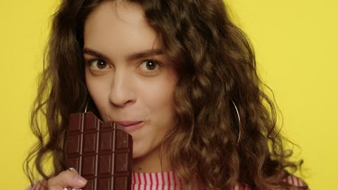 Happy woman enjoy chocolate bar in slow motion. Model girl with chocolate posing on camera on yellow background. Portrait of young woman eating dark chocolate bar