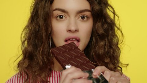 Young woman opening chocolate bar in studio. Portrait of happy girl eating chocolate in slow motion. Portrait of fashion model eating chocolate bar on yellow. Close up of pretty girl eat sweet food