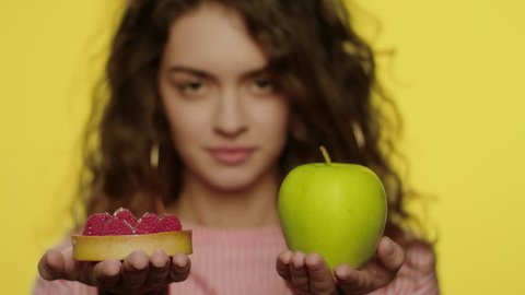 Young woman choosing apple instead cake for diet nutrition in studio. Portrait of cheerful girl choosing fruit diet for healthy nutrition and weight loss. Healthy food concept