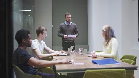 Tracking shot of business team sitting at meeting table in boardroom and discussing financial strategy behind transparent glass wall. Manager speaking and asking questions, teammates taking notes