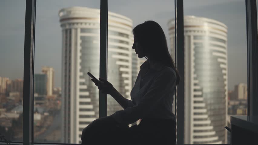 Silhouette of a girl with a phone in her hands on the background of modern buildings. business woman uses a smartphone in the office. | Shutterstock HD Video #1026992162