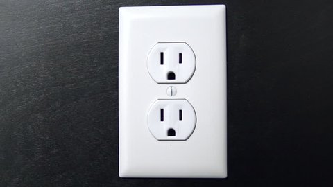 Male hand plugs in and unplugs a two pronged cord into the bottom outlet of an American electrical socket, centered close up