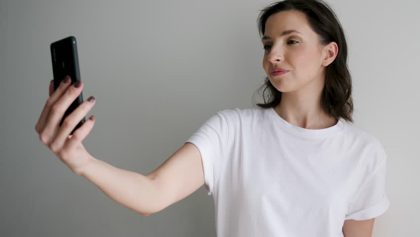girl in a white t-shirt on a light wall makes a selfie on a black phone by the window