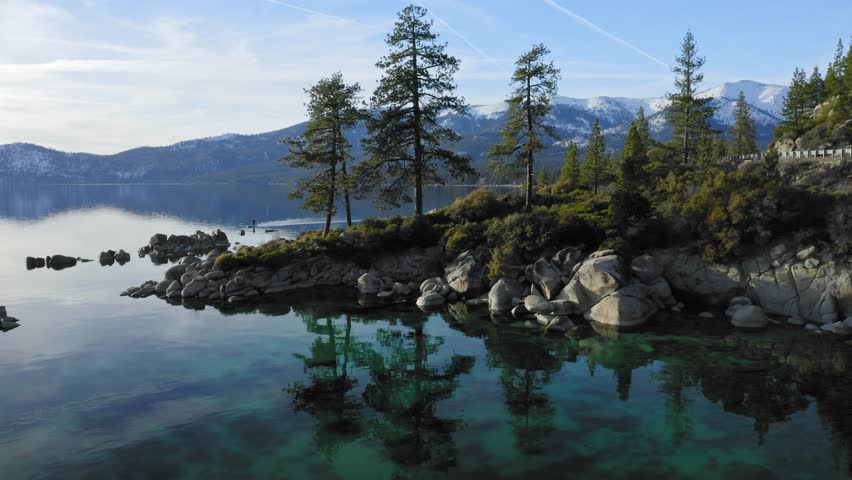 Aerial: flying over a flat mirror lake, rocks pine trees on the foreshore in Lake Tahoe. In the background is mountains covered in snow. Nevada, USA 