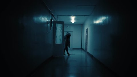 Tracking inside a long dark gloomy corridor. A dark silhouette of a girl dressed in a parka stops in front of the camera.White big clocks hang on the wall and show ten to eleven.Concept of horror.