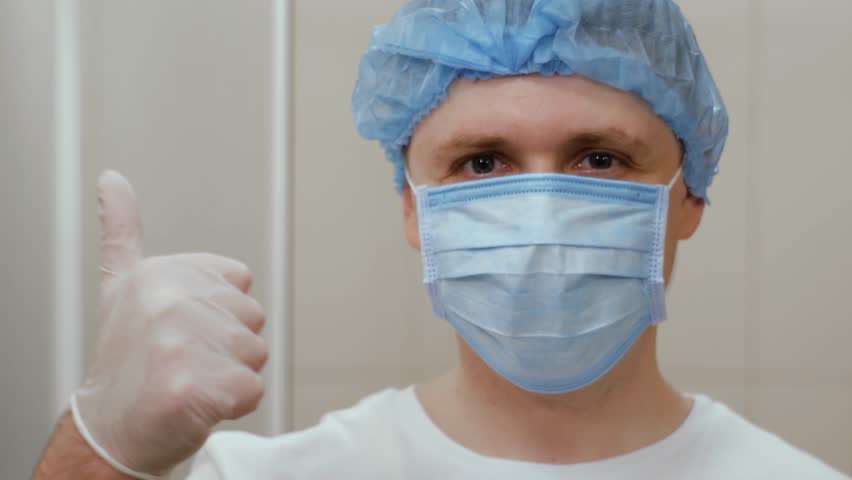 Happy doctor nods his head approvingly and shows thumb up. Man in a medical mask looks straight ahead and gives approval