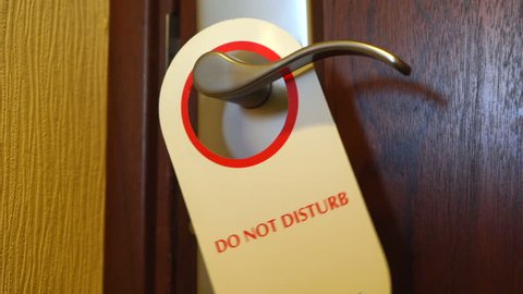 Do Not Disturb sign swinging on hotel room door. Red white door hanger on handle close-up.
