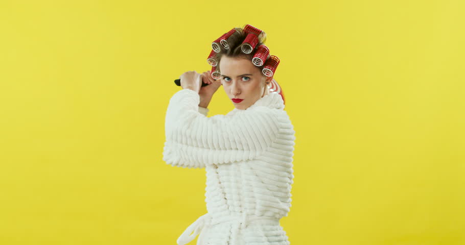 Caucasian pretty young girl in the white bathrobe and curlers holding a frying pan and swinging it as want to beat someone on the yellow wall back.