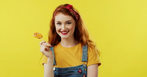 Portrait shot of the young Caucasian pretty girl with red hair standing on the yellow screen background, holding a lollipop and smiling joyfully. Close up.