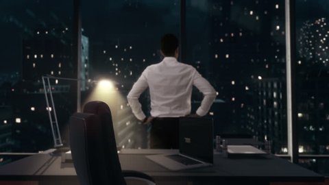 Back view of thoughtful Businessman standing in his office working late at night, looking out of the window as it rains contemplating his next big idea. Ultra HD 4K.