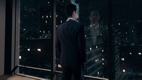 Thoughtful Businessman wearing a suit standing in his office on a rainy night, looking out of the window and contemplating his next big business deal.