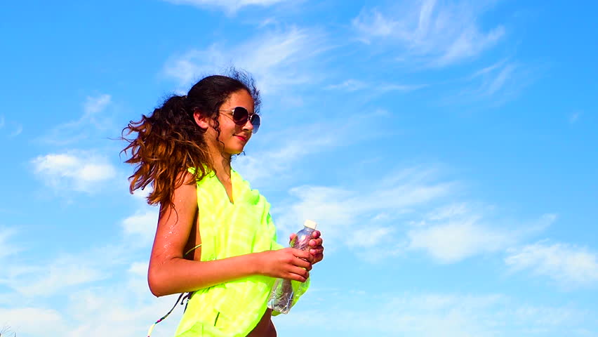 Side view of a beautiful girl in a bathing suit sitting on the sand drinking water after a workout. Slow motion