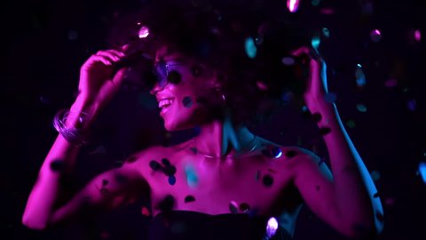 Young african girl with curly hair dancing and having fun in confetti rain on black background. Woman celebrating, depicts joy and happiness. Success, party, holiday concept