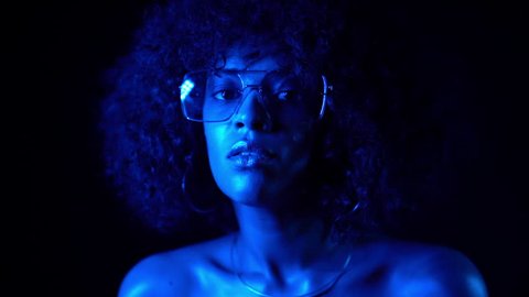 Disco sexy woman with transparent sunglasses posing in dark room at night in blue neon light. Portrait of young mixed race african girl. Fashion, glamour, model concept