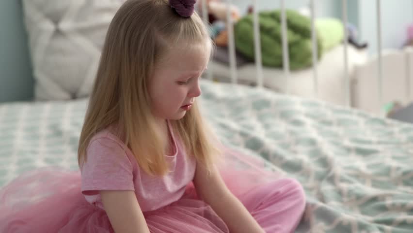The little girl in the pink dress is very angry and upset, frowning her brow and showing tongue. Bad mannered little girl with blond hair. Crying on the bed | Shutterstock HD Video #1027079411