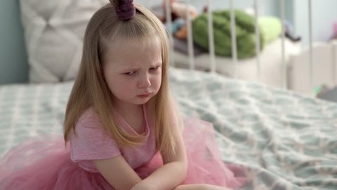 The little girl in the pink dress is very angry and upset, frowning her brow and showing tongue. Bad mannered little girl with blond hair. Crying on the bed