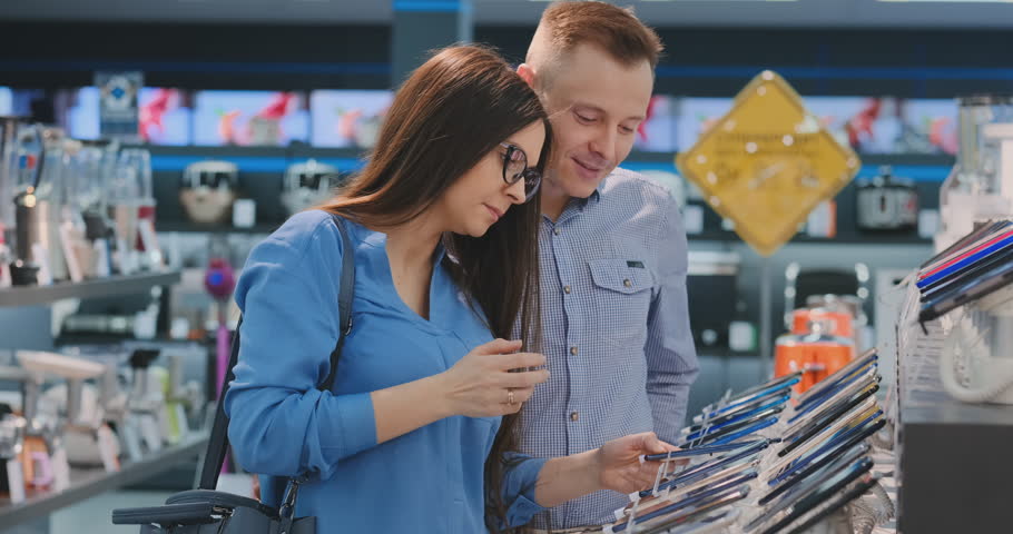 Young beautiful married couple a man in a shirt and a woman in glasses holding a smartphone standing around a showcase with smartphones in an electronics store checking the touchscreen of the screen Royalty-Free Stock Footage #1027085609