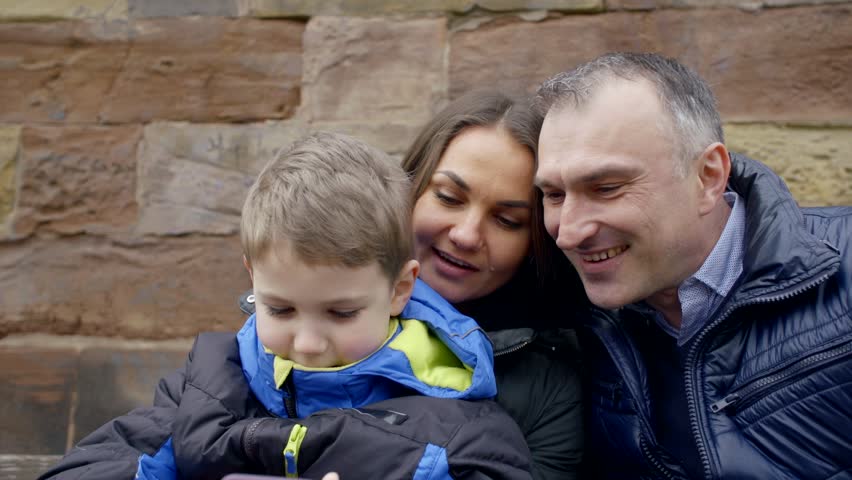 child boy is playing smartphone walking with parents outdoor, mother is holding his