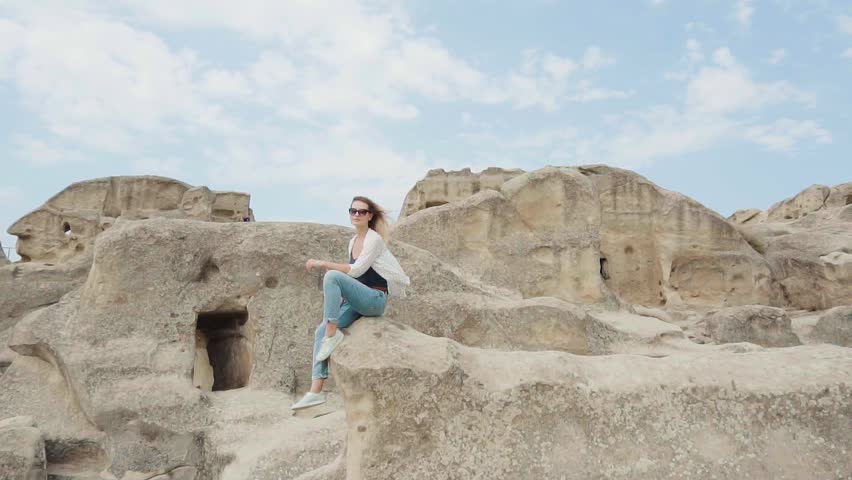 pretty young woman sits and rests on small stone ledge like on rock, lady with bright flying hair admires magnificent view from mountains against bright cloudy sky, attractive tourist as philosopher