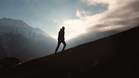Very long shot Aerial view of epic shot of a man walking on the edge of the mountain as a silhouette in a beautiful sunset. Silhouette of a man with a beard and wearing a hat climbing uphill