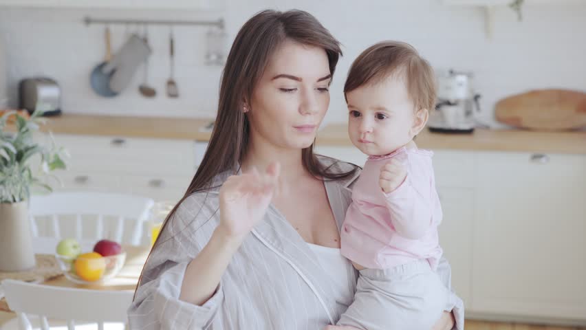 Young mother holding baby daughter in arms and teaching her how to wave goodbye to guests and talking to her