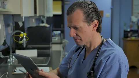 Portrait of a male nurse, doctor, or surgeon working on a digital tablet in a hospital or medical office looking up and smiling at the camera in slow motion