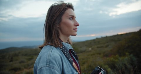 Outdoor spring smiling lifestyle shot of pretty young woman taking photos of sunset in the mountains in Europe, travel photographer making pictures at evening, slow motion, cine camera 4K
