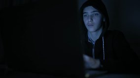 Hacker's hacking into the computer network. A young guy in a dark room typing on a laptop.  Hacker with a laptop. Lonely teen chatting online. Computer criminal.