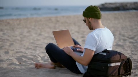 Young urban digital nomad of millennial generation z age sits on beach at resort or travel vacation destination, works on laptop, updates application or checks email notifications, remote office job