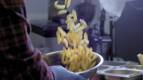 The cook lowers into a boiling oil fryer with French fries. Metal box can for raw potato. The kitchen of a fast food restaurant. Making chips. Raw potato chips in stainless bowl