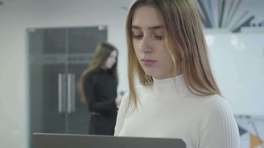 Portrait of the pretty lady searching for information in her netbook at work. Attractive woman gives high five to older bearded man just entering the room. Morning time in the office