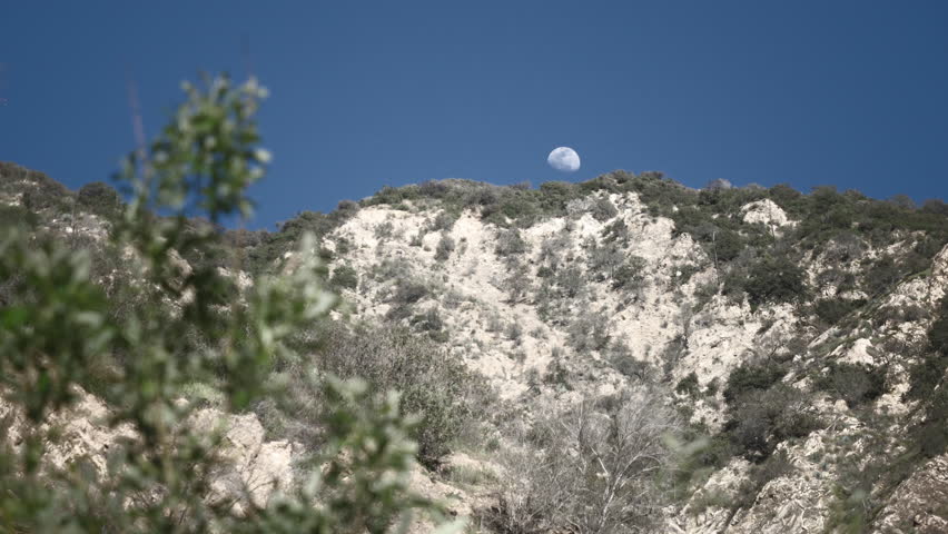 Moon and mountain with blue sky. Rocky Mountains with half moon background at day. Elegant Moon during a day light. Stunning full moon rises above mountains edge in canyon. Creepy ashes mountains.