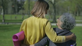 Rear view of young woman in yellow sweatshirt and middle-aged woman with grey hair walking in park, embracing each other, holding yoga mat in hands, laughing. Sport, communication concept