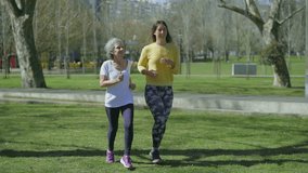 Smiling young woman in yellow sweatshirt and excited middle-aged woman with grey hair in white T-shirt jogging in park, talking, looking happy. Sport concept