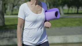 Medium shot of middle-aged Caucasian female body in white T-shirt walking in park, holding yoga mat in hand. Sport, lifestyle concept