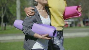 Medium shot of bodies of young woman in yellow sweatshirt and middle-aged woman with grey hair walking in park, embracing each other, holding yoga mat in hands, laughing. Sport, communication concept