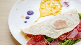 Footage of delicious tortilla with jamon ham slice,fresh spinach and cherry tomato served on white plate in cafe for lunch.Video of piadina dish in close up
