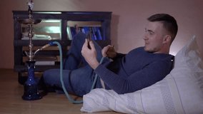 A young guy smokes a hookah, exhales smoke and talks on video by phone. A young man in the smoke. A young guy on a pillow against the background of a bookcase and holds a phone in his hands.