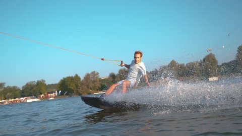 SUPER SLOW MOTION, CLOSE UP: Glassy drops of water fly at the camera as the joyful fit man wakesurfs on the calm lake during a fun summer vacation. Athletic tourist smiles as he wakesurfs past camera.