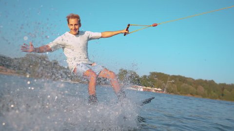 SLOW MOTION CLOSE UP: Fit young Caucasian man smiles as he wakesurfs on the tranquil lake and splashes the water at the camera. Refreshing drops of water fly at the camera after wakesurfer speeds past