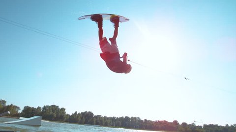 SLOW MOTION LENS FLARE: Wakesurfer does a spinning barrel roll while riding around the lake on a sunny summer day. Bright sunbeams shine on the athletic man doing tricks in the wakeboarding water park